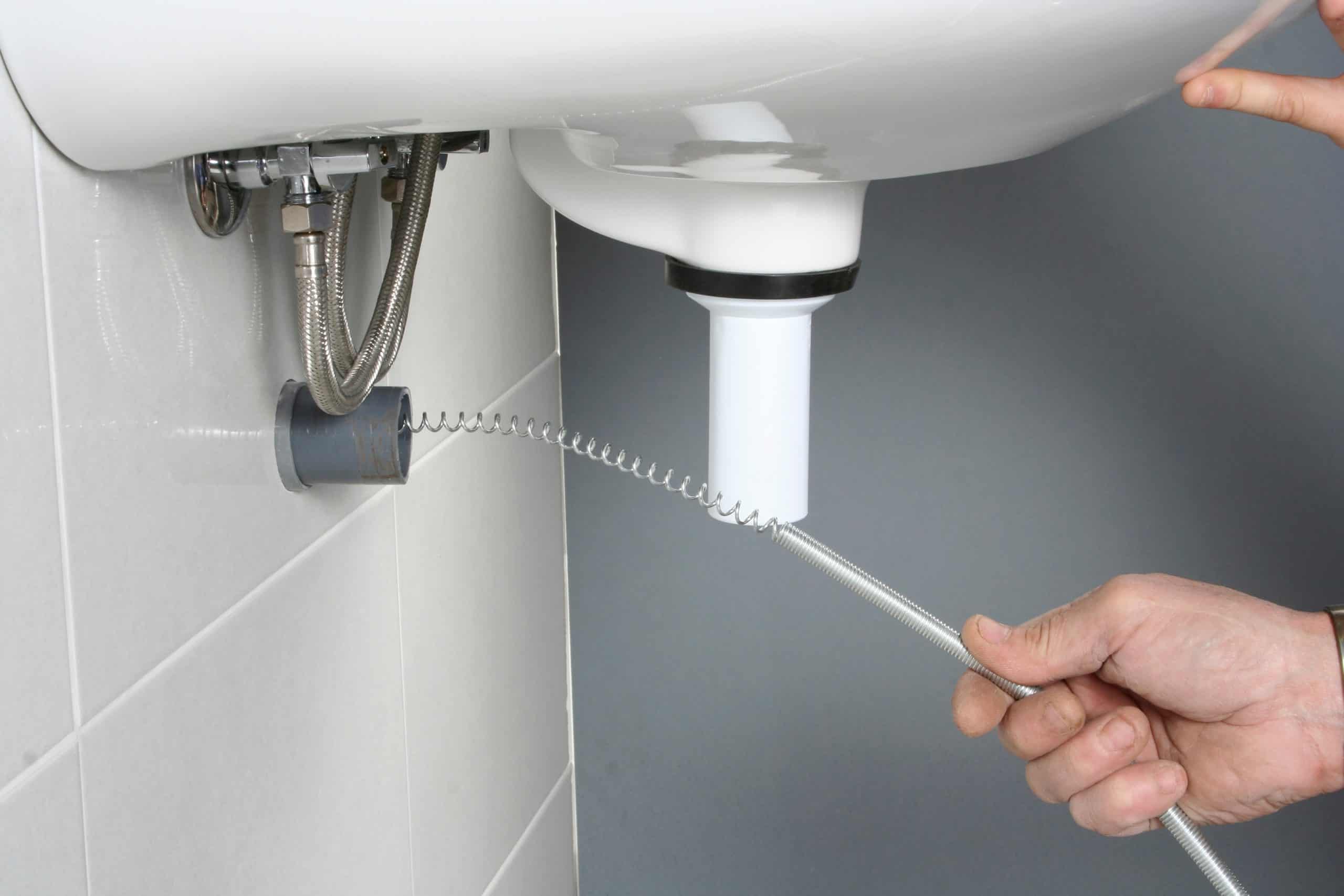 A person using a plumbing snake to clear a blockage in a white sink drain. The hand is holding the tool under the sink, which is fixed to a tiled wall. A pipe and other plumbing fixtures are visible.