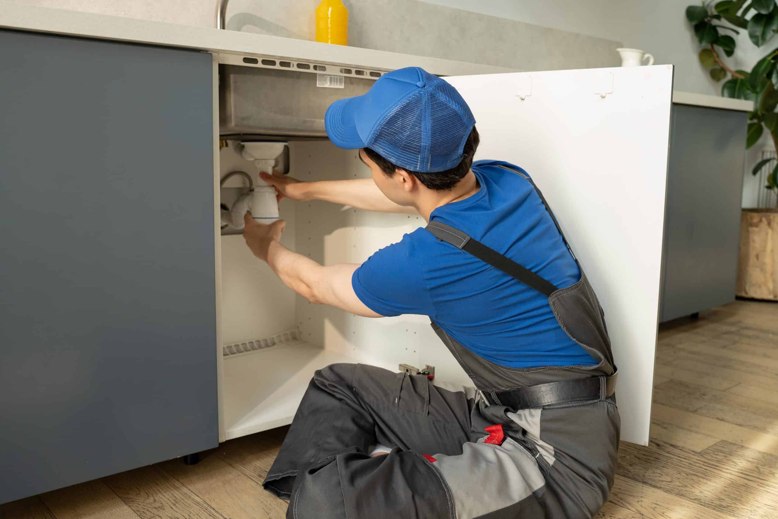 A plumber wearing a blue cap and shirt works under a kitchen sink, inspecting the plumbing. The kitchen has dark blue cabinets and a wooden floor, with a plant in the corner and a yellow bottle on the counter.