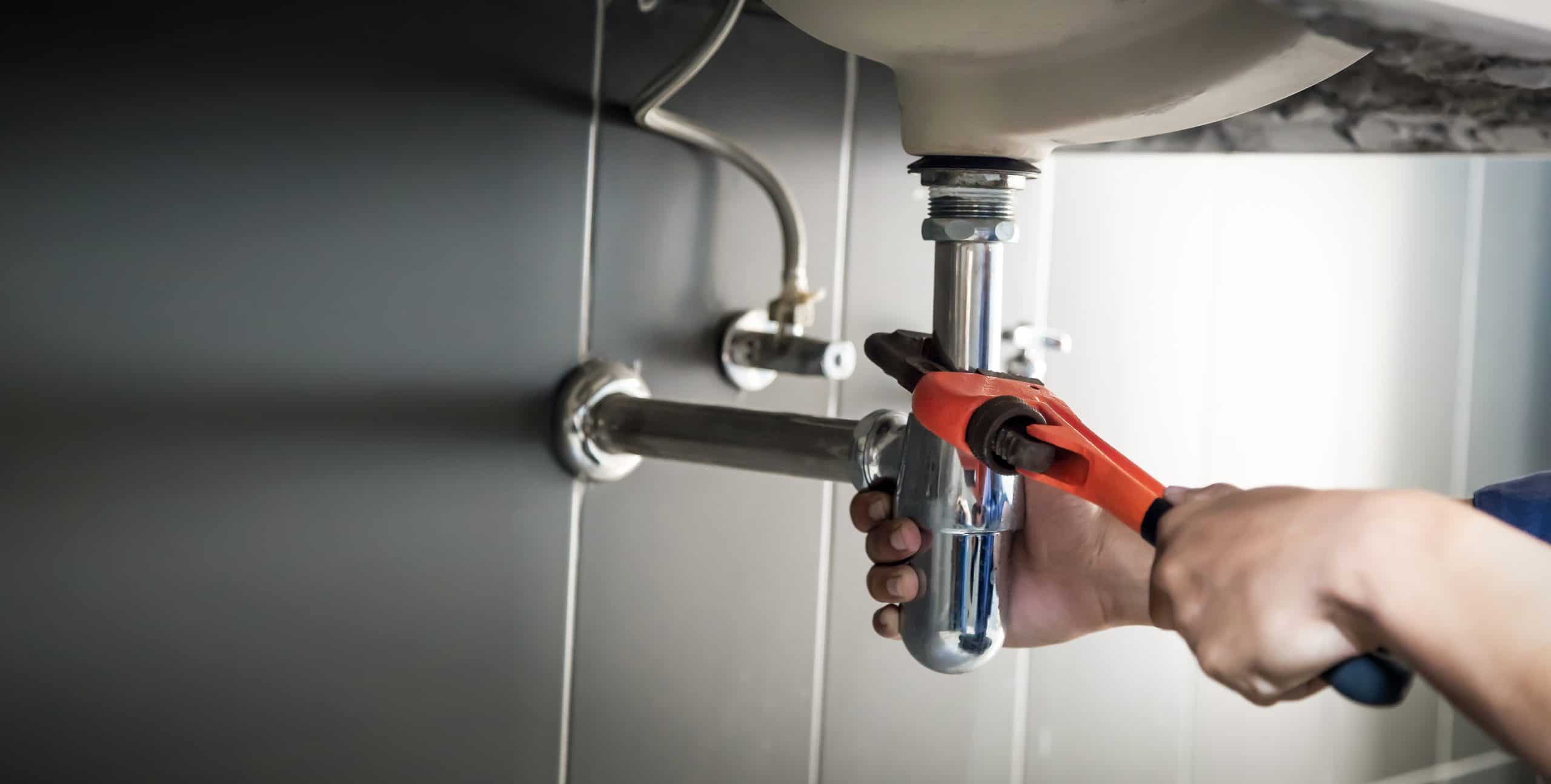 A person uses a red pipe wrench to tighten plumbing under a sink. The hands grip the shiny metal pipe, which connects to the sink's drain. The background shows tiled walls and additional plumbing components.