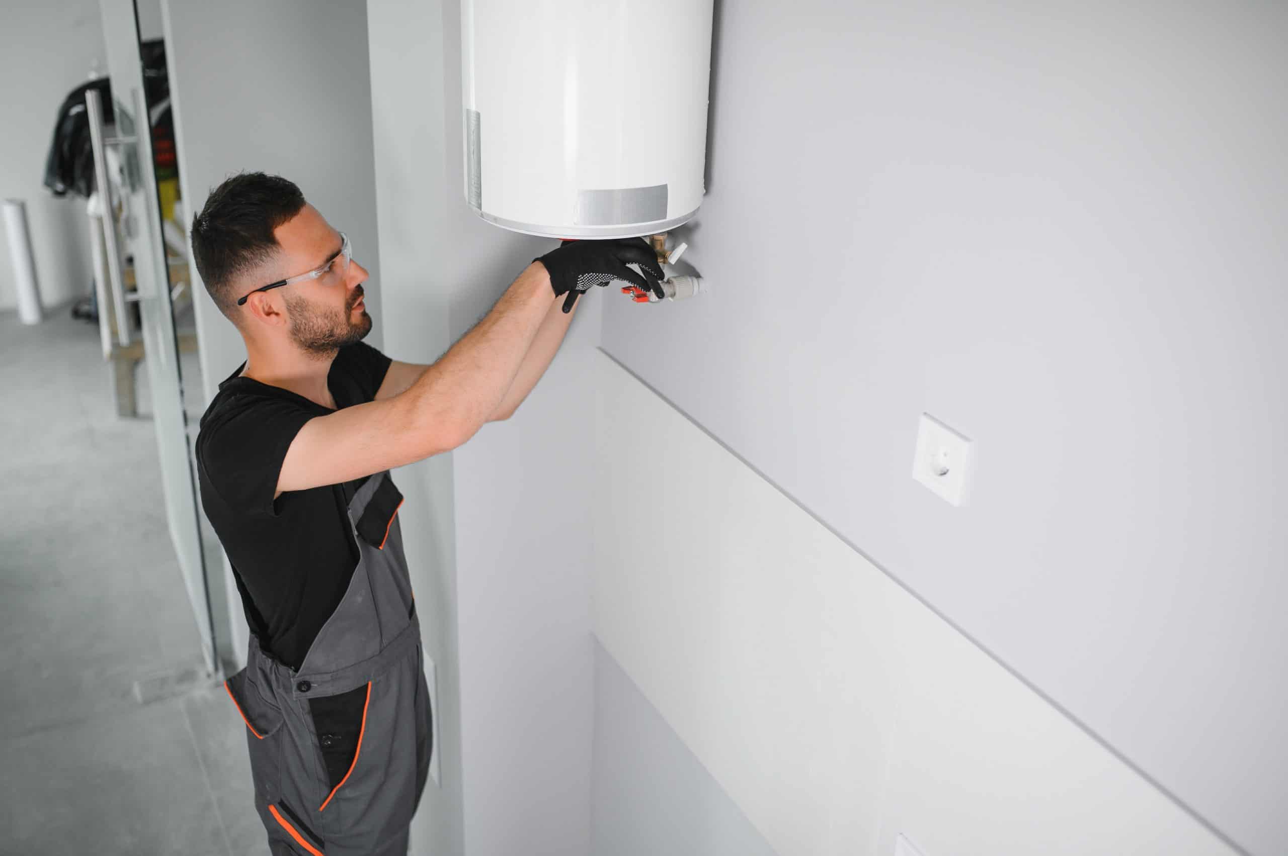 A man wearing glasses and work gloves is installing or repairing a white wall-mounted appliance, possibly a heater, in a modern, minimalistic room. He's dressed in a black shirt and dark overalls.