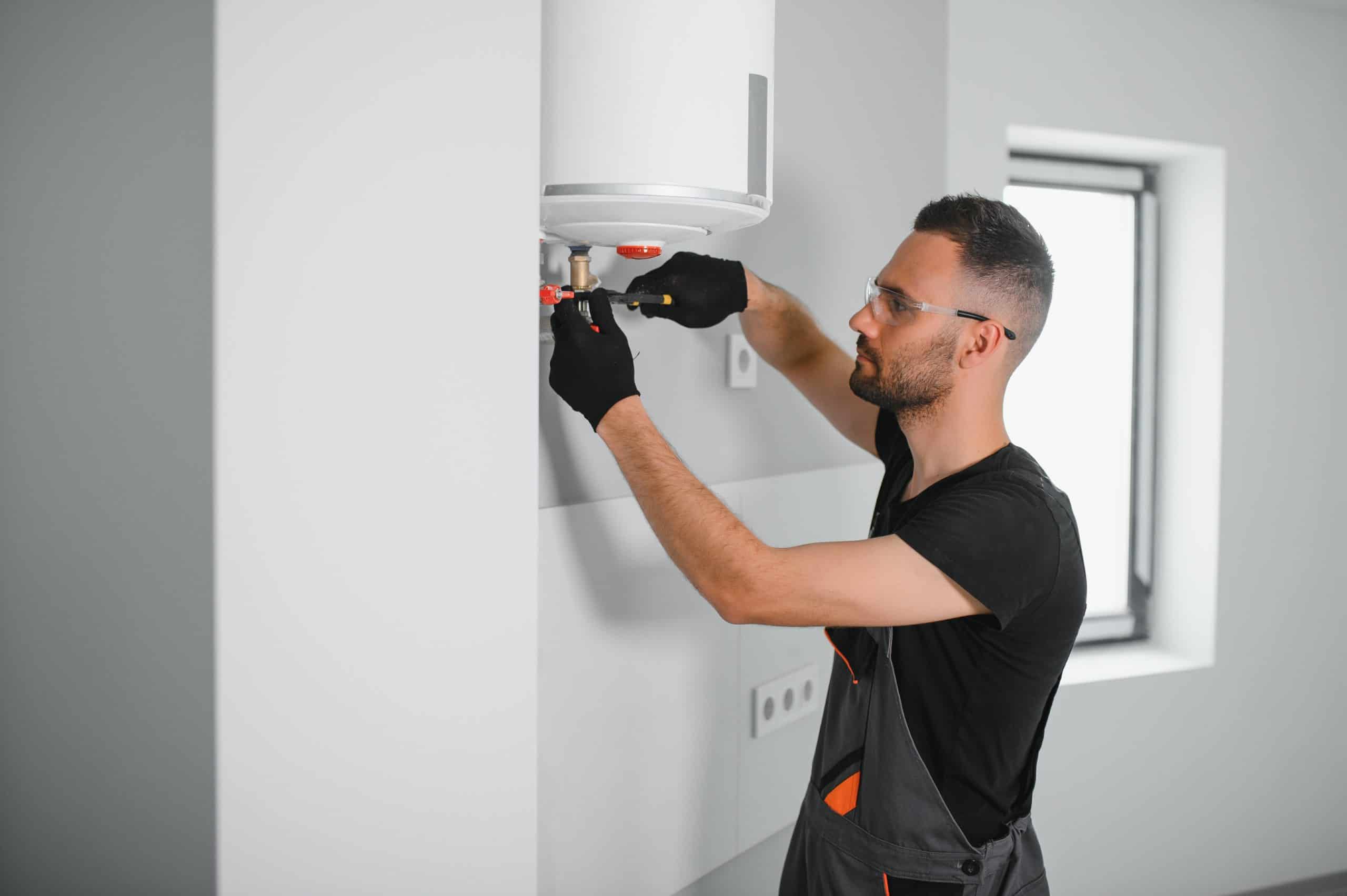A man wearing black gloves and safety glasses is installing or repairing a white water heater on a wall. He is focused on adjusting a component with tools. The room is modern with a window in the background.