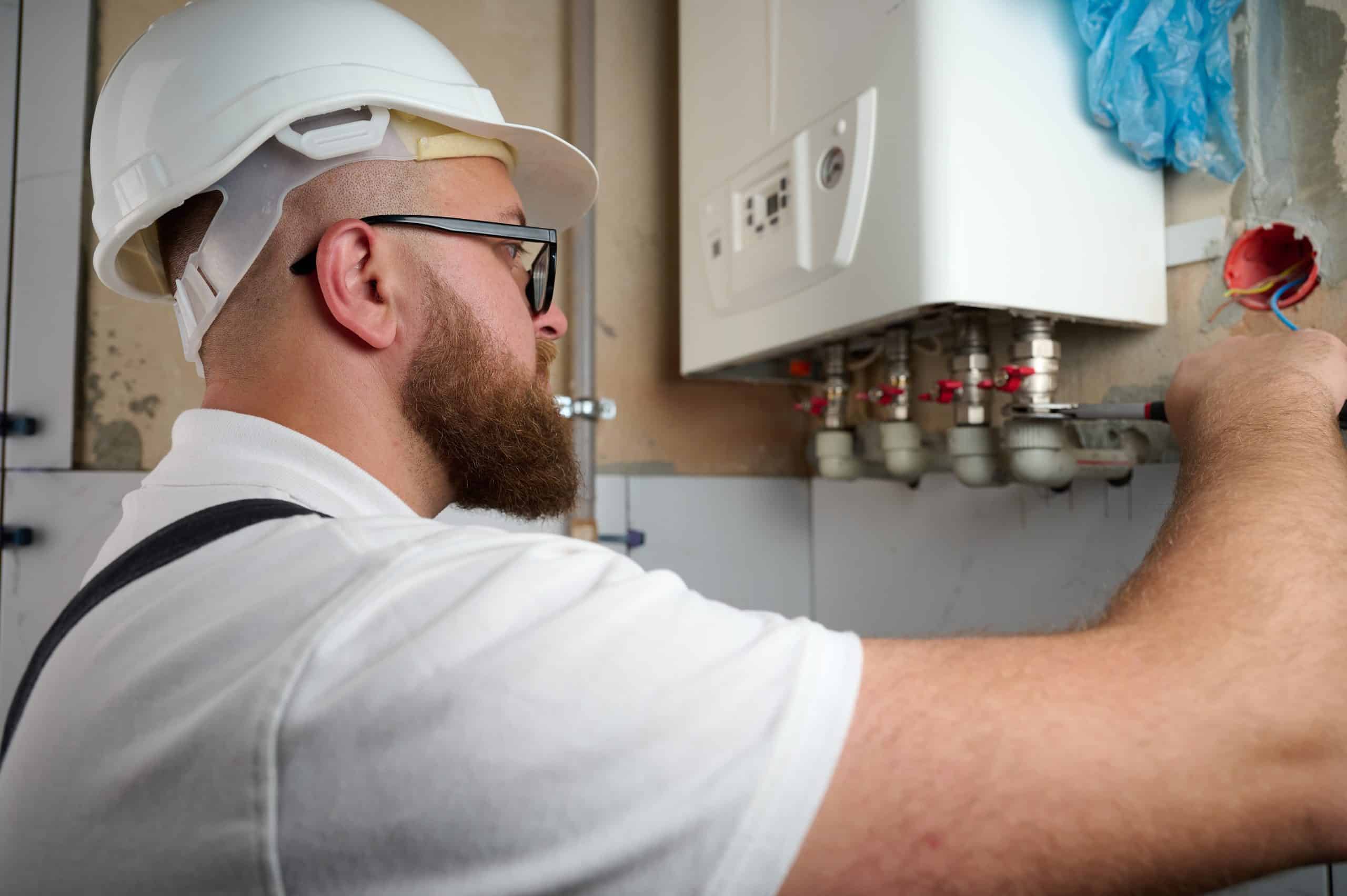 A bearded technician in a white hard hat and glasses works on a boiler, adjusting pipes with a wrench. He wears a white shirt and black suspenders, concentrating on the task in a workshop setting.