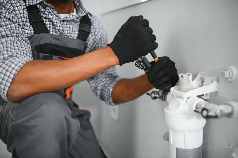 A person wearing black gloves and a checkered shirt is using a wrench to work on plumbing pipes connected to a white filter. They are crouched down, focused on the task in a light-colored room.