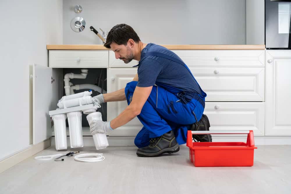 A plumber in blue overalls installs a water filtration system under a kitchen sink. The white cabinetry contrasts with the red toolbox on the floor. Various plumbing tools are scattered nearby.