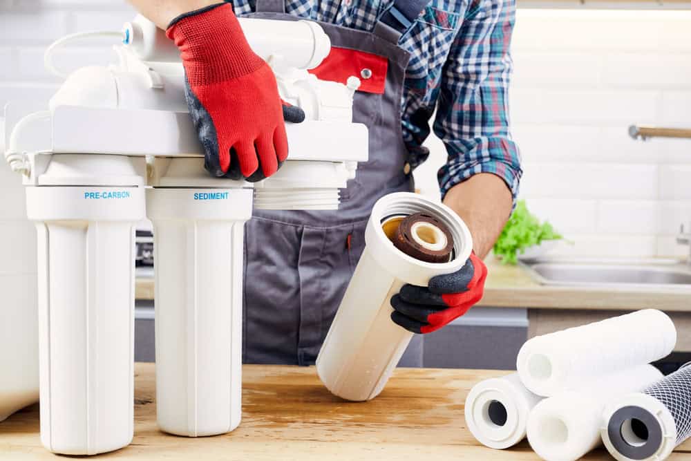 A person wearing red gloves is replacing a water filter in a modern kitchen. Various filter cartridges are scattered on the counter. The individual is dressed in a checkered shirt and grey overalls, focusing on the task at hand.